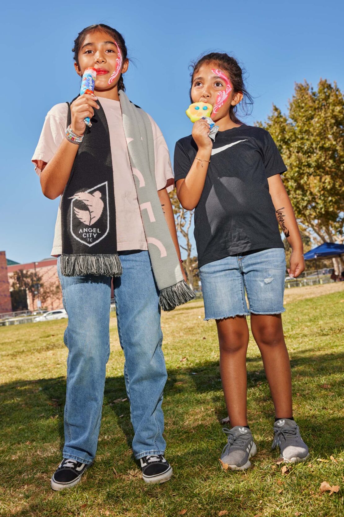 Two smiling girls with face paint are enjoying popsicles outdoors, one wears an Angel City scarf; grass, trees and blue sky in background.