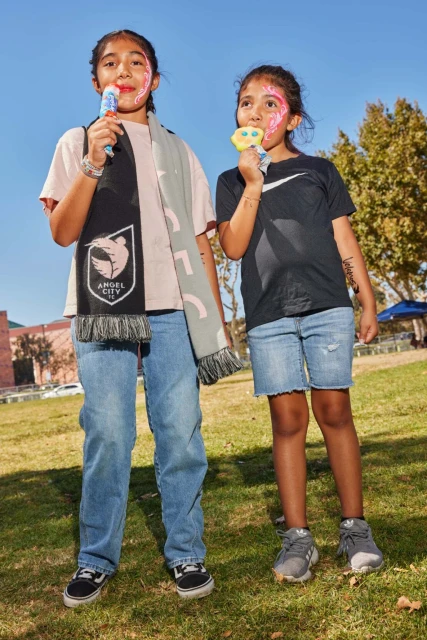Two smiling girls with face paint are enjoying popsicles outdoors, one wears an Angel City scarf; grass, trees and blue sky in background.
