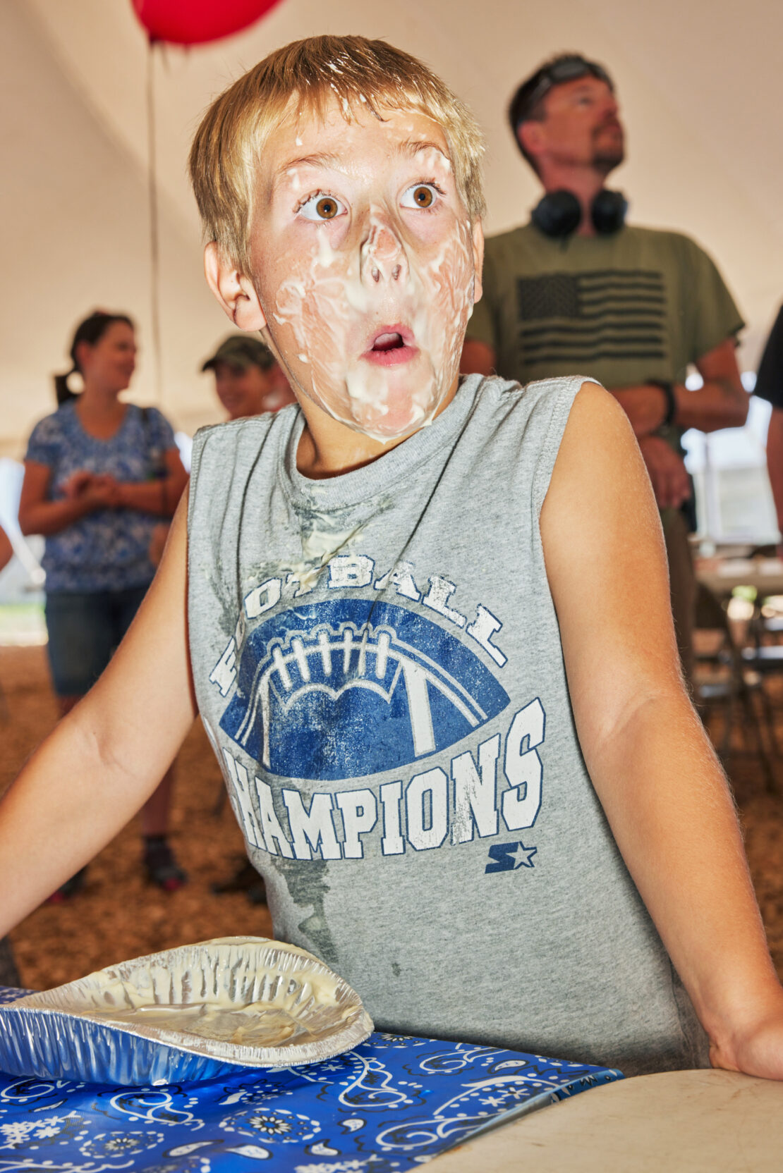 A young boy with cream all over his face reacts with surprise, standing next to a table with a patterned tablecloth, indoors.
