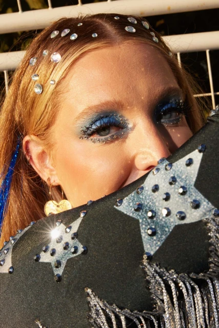 Young woman with blue eyeshadow and rhinestone embellishments, partially obscured by a black fringed hat, close-up studio shot.