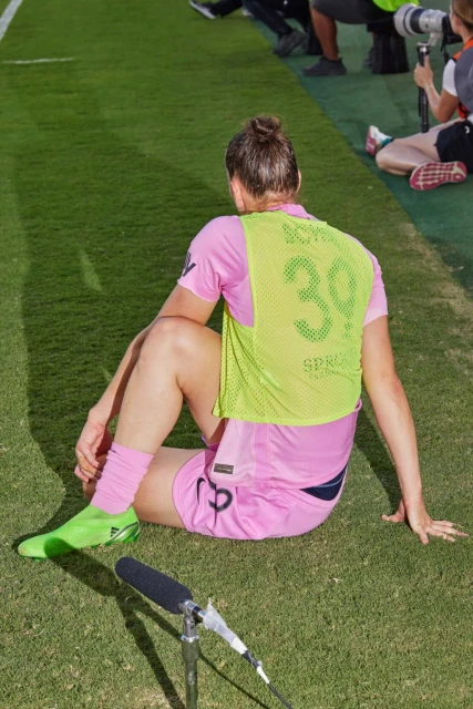 Female soccer player rests on the field wearing a pink jersey and green cleats, with field markings and a microphone nearby.