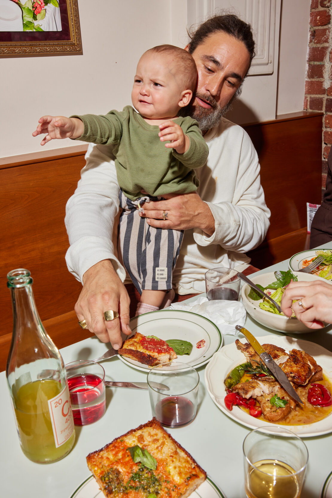 A father holds his baby at a restaurant table with plates of food and drinks, creating a warm, family-oriented setting with indoor lighting.