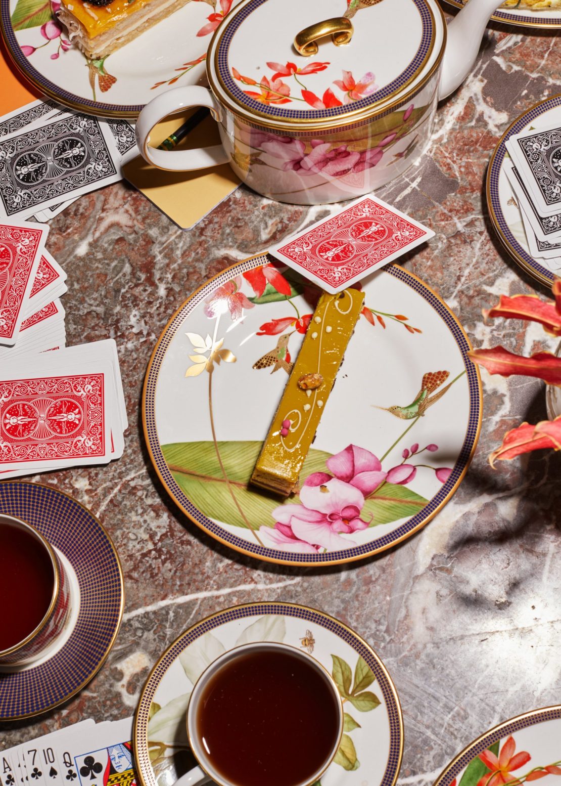 Overhead shot of floral tea set, playing cards, and dessert on a marble table. Warm lighting enhances the rich colors and intricate details.
