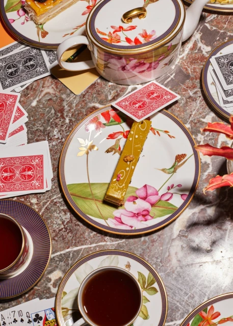 Overhead shot of floral tea set, playing cards, and dessert on a marble table. Warm lighting enhances the rich colors and intricate details.