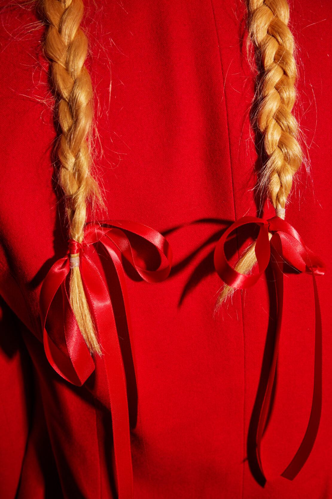 Close-up of blonde braids tied with red ribbon against a vibrant red background, creating a striking visual contrast.