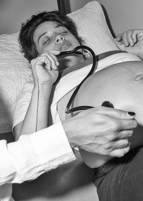 Monochrome image of a pregnant woman lying in bed while a healthcare provider listens to her baby's heartbeat with a stethoscope.