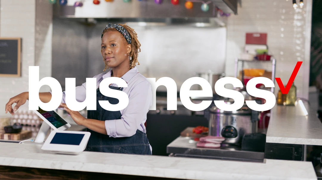 An adult Black woman wearing a floral headscarf uses a touch-screen POS system in a restaurant with professional kitchen equipment in the background.