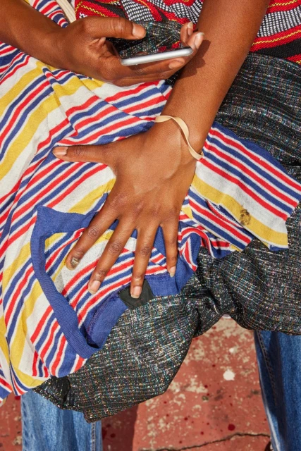 Close-up shot of a person of color holding a mobile phone. They're wearing colorful, striped fabric and blue jeans in outdoor lighting.