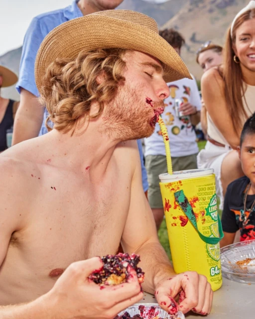 A person with a straw hat is eating a messy berry pie outdoors with other people in the background on a sunny day.