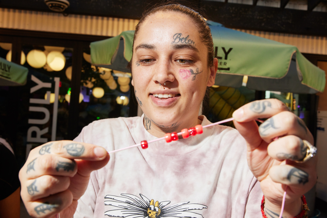 Adult female with face and hand tattoos smiles and holds beaded necklace; wearing a daisy t-shirt in an indoor setting with a Truly Hard Seltzer sign.