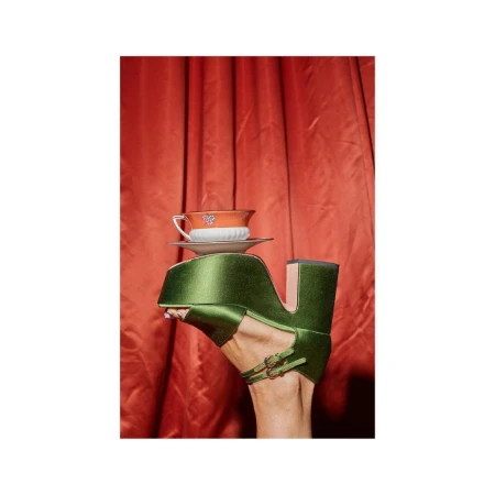 Green platform shoe supports a teacup on a foot with an orange curtain background, creating a whimsical fashion-forward visual.