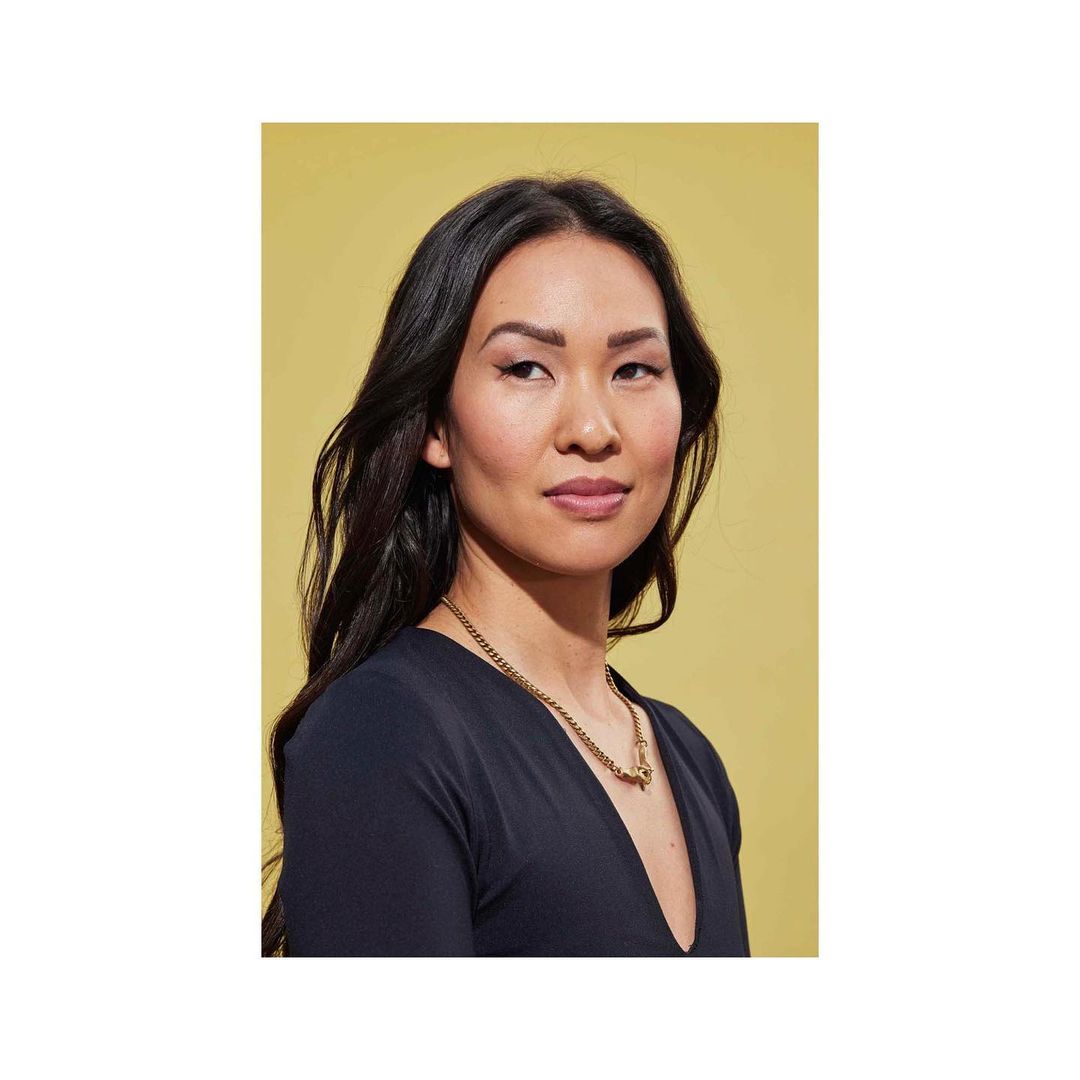 Portrait of a woman with long dark hair wearing a gold chain necklace and dark top against a yellow background