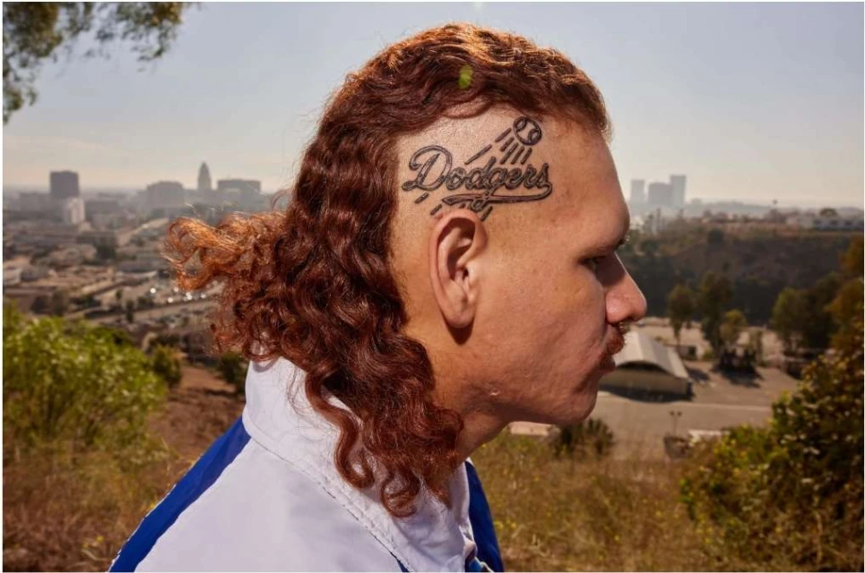 Outdoor portrait of an adult male with curly hair and a Dodgers tattoo, with a city skyline visible in the background on a sunny day.