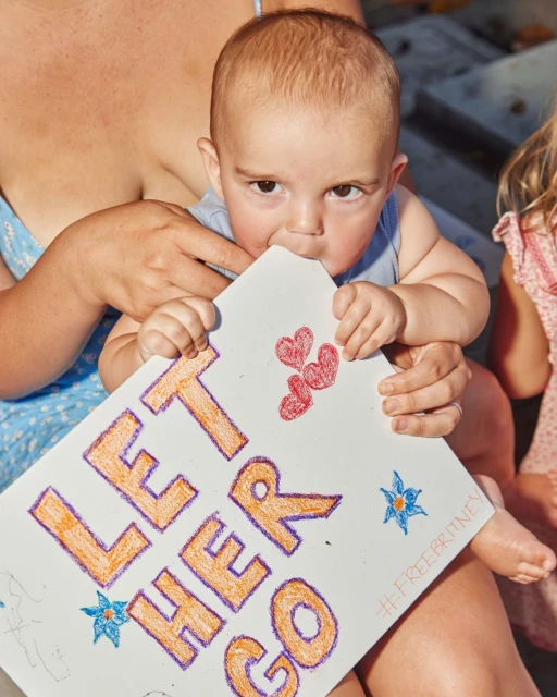 A baby is sitting on someone's lap, holding a sign with the words 'LET HER GO' drawn on it, adorned with hearts and star doodles.