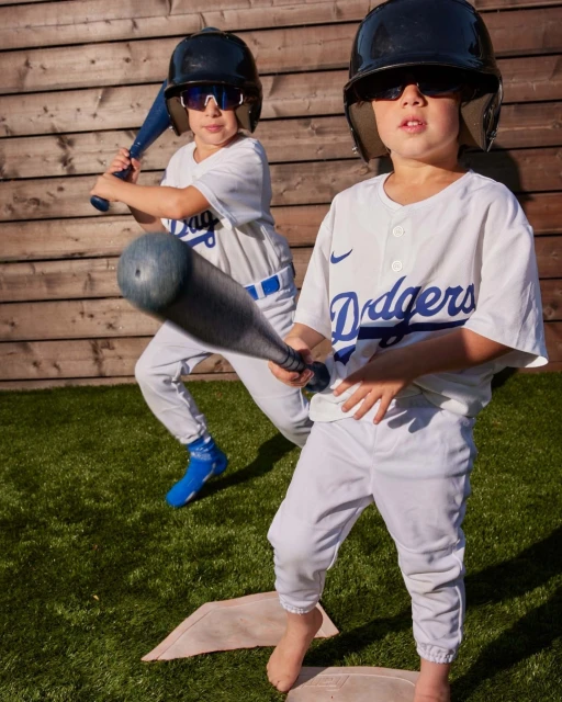 Two young boys dressed in baseball uniforms and helmets are holding bats on a baseball field with a wooden fence in the background.