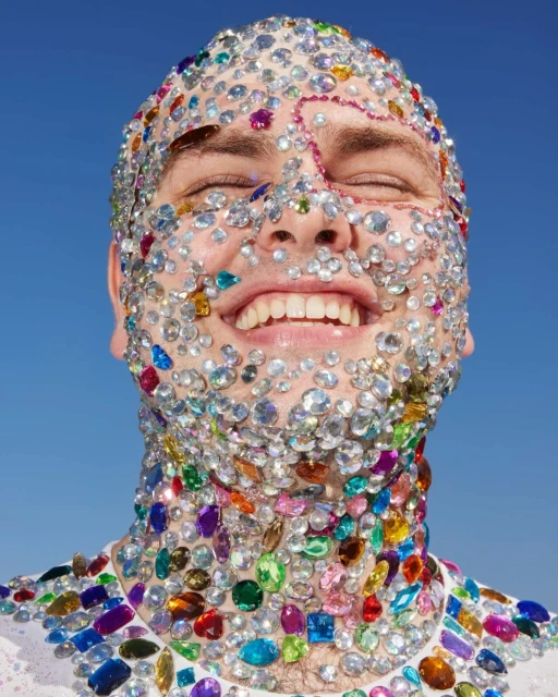 Adult male smiles with eyes closed, face and torso covered in large colorful rhinestones against a bright blue sky background.
