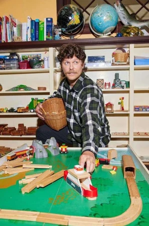 Man sits at a toy train table holding a woven basket, surrounded by wooden train tracks and toy trains with a bookshelf background.