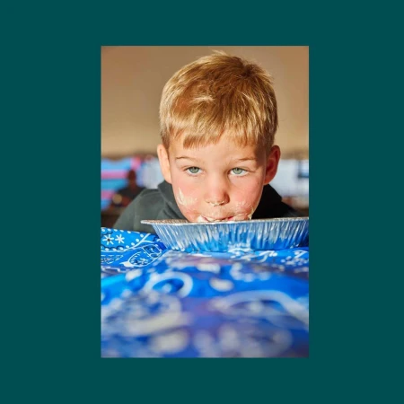 A young blond boy with food on his face is about to eat from a pie tin with a blue bandana-patterned tablecloth underneath the pie.