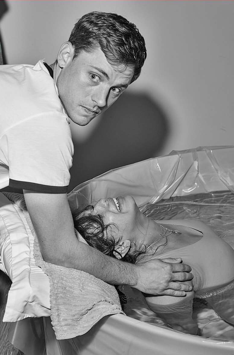 Monochromatic shot of a man assisting a woman in a birthing tub, featuring water, pillows, and an intimate setting. The woman smiles.