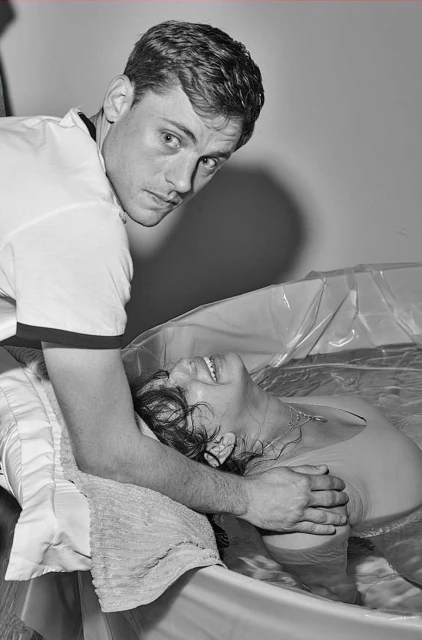 Monochromatic shot of a man assisting a woman in a birthing tub, featuring water, pillows, and an intimate setting. The woman smiles.