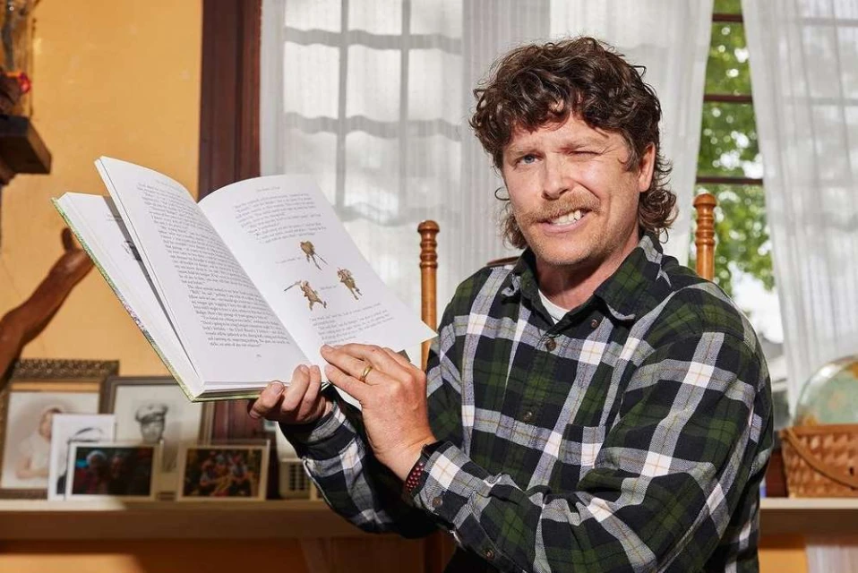 Adult male reading a book with illustrations indoors. He is wearing a plaid shirt and smiles at the camera while winking one eye.