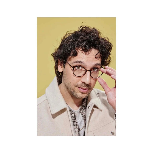 Adult male poses indoors wearing glasses, tan jacket, and patterned shirt against a yellow background in a studio portrait.