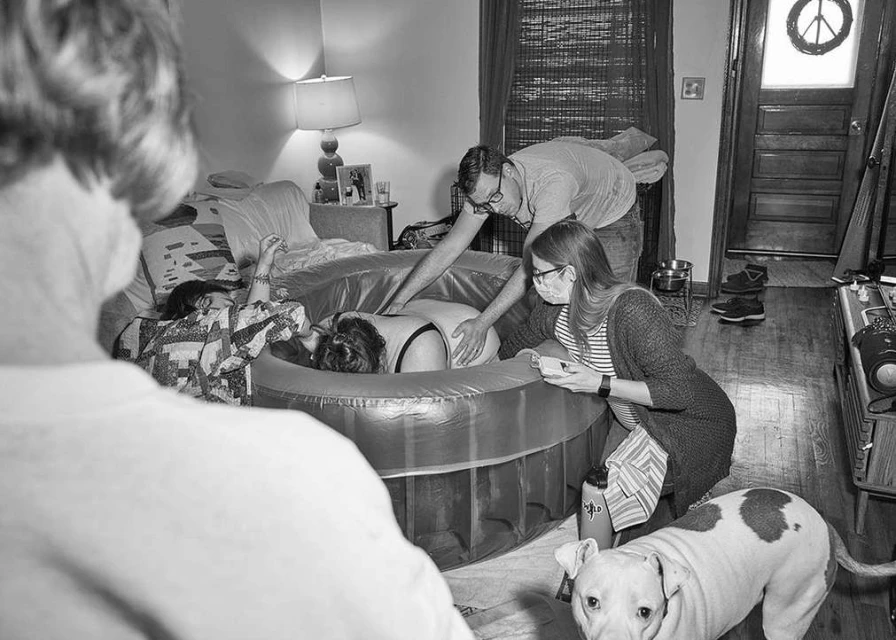Black and white photo of a woman in a birthing pool with two adults assisting, one child, and a dog in a domestic setting.