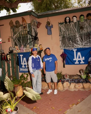 Two men stand in front of a house adorned with Halloween decorations, including masks, cobwebs, and a Dodgers banner. A large potted plant.