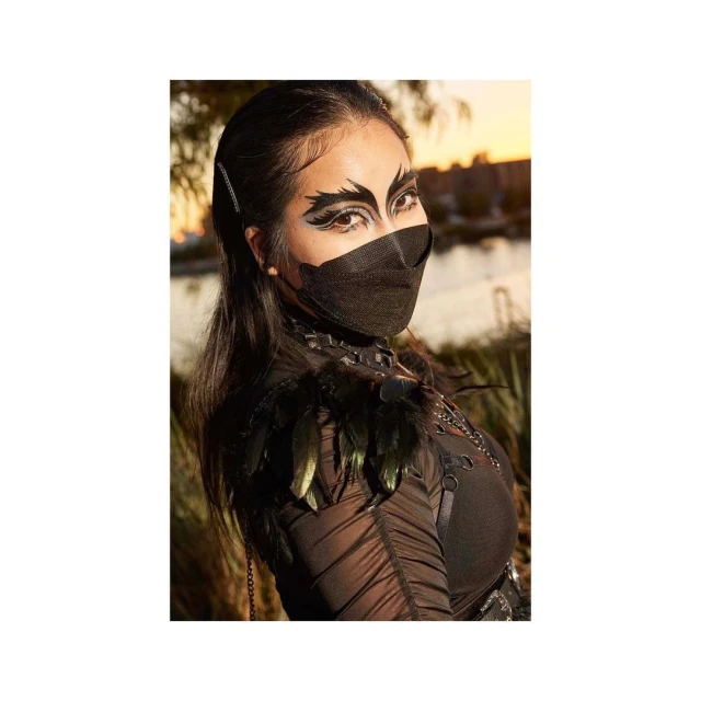 Portrait of adult female in black costume and mask with theatrical eye makeup in outdoor environment with neutral colors in background.