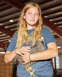 Young girl holding a barred Plymouth Rock chicken in an agricultural shed interior by photographer Maggie Shannon
