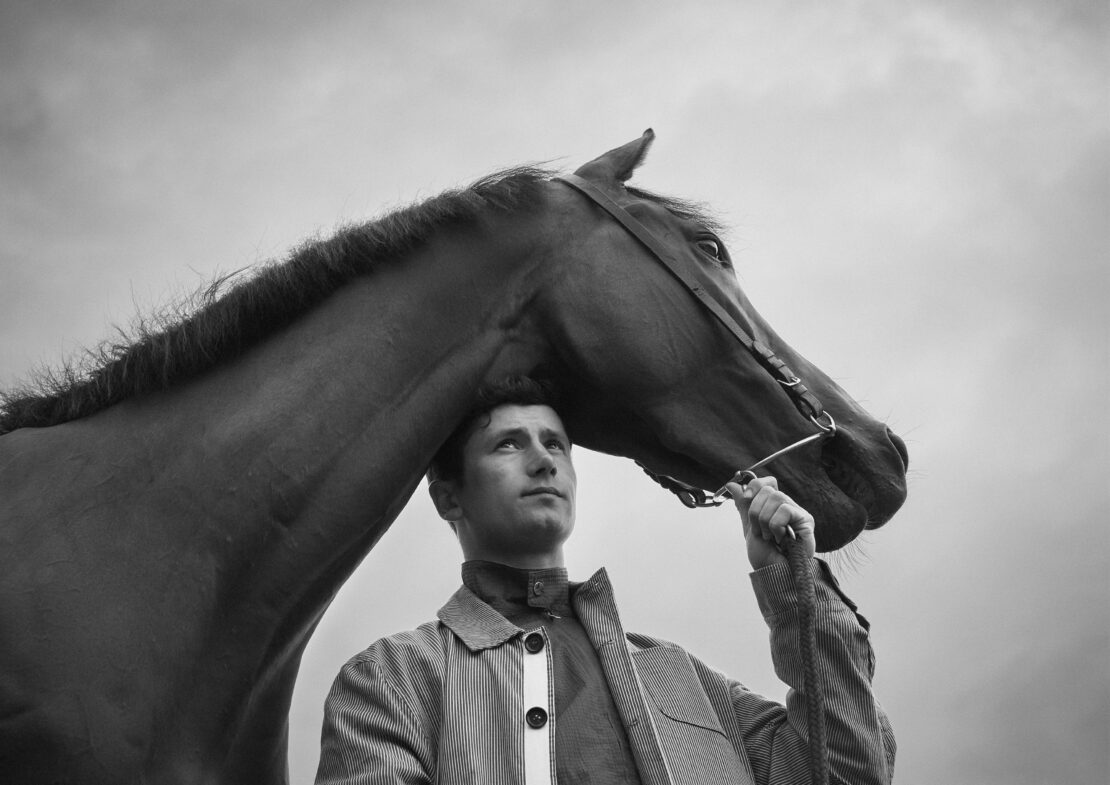 Monochrome portrait of a horse and adult male outdoors, featuring a pinstripe coat. The horse's head is near the man's shoulder.