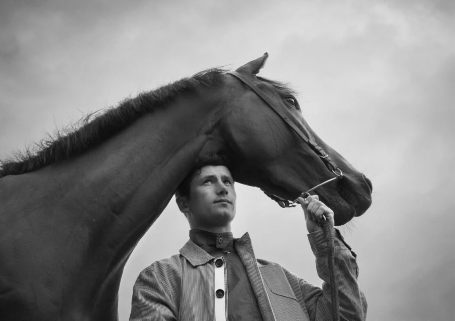 Monochrome portrait of a horse and adult male outdoors, featuring a pinstripe coat. The horse's head is near the man's shoulder.