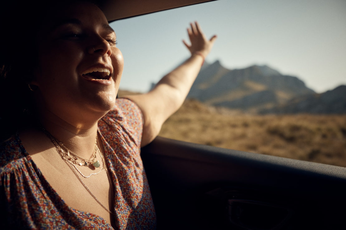 A woman with open mouth and raised arm enjoys the wind and sunlight during a car trip, with mountains visible in the background.