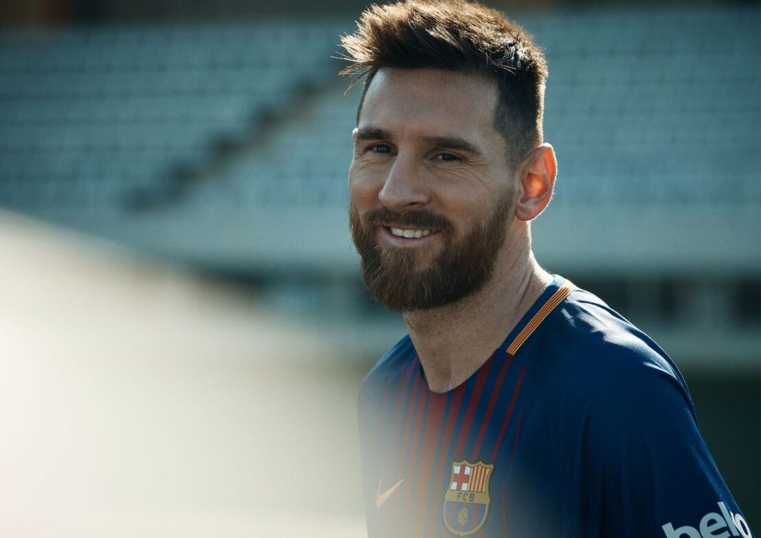 Smiling soccer player in FC Barcelona jersey stands on the field, looking confidently at the camera with blurred stadium background.