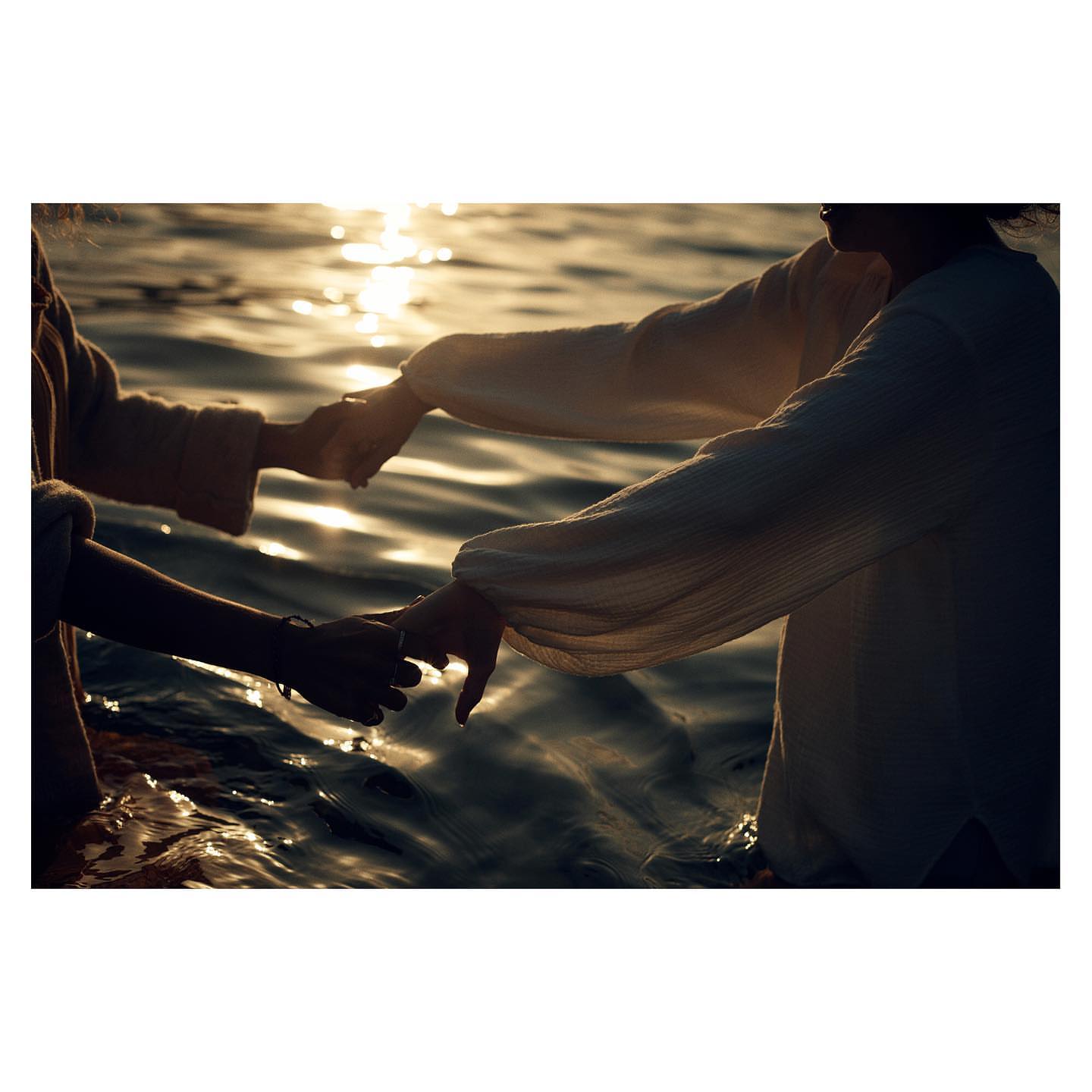 Two people holding hands while standing waist-deep in water at sunset, captured in silhouette.