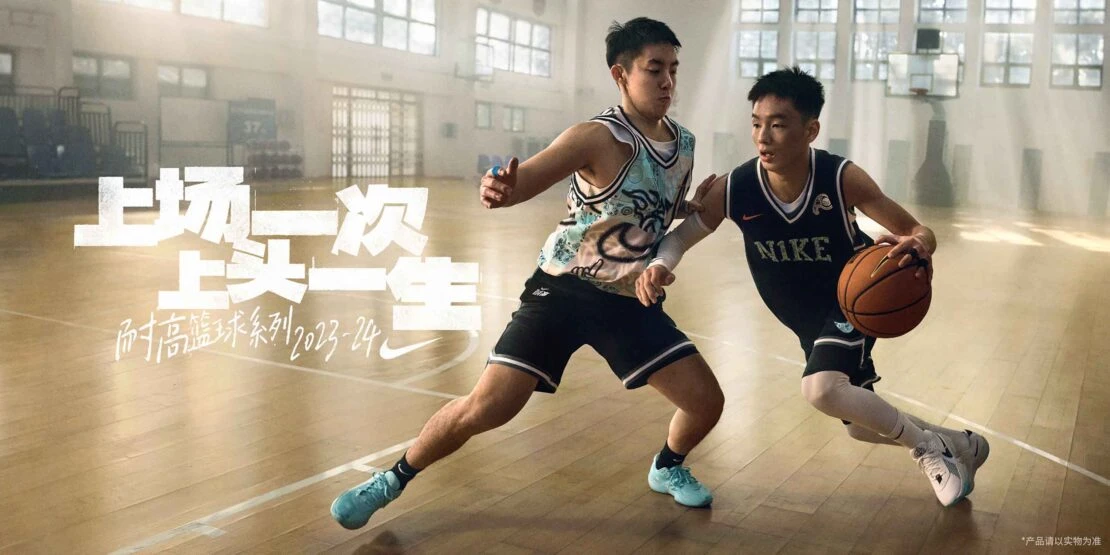 Two basketball players dribbling in a gymnasium with natural light, wearing Nike uniforms, athletic shoes. Promotional image.
