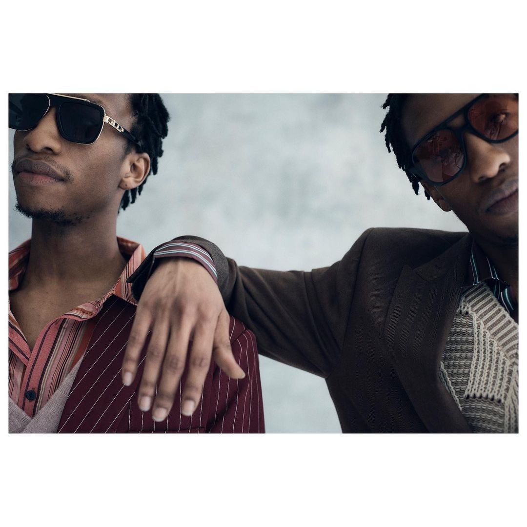 Two stylish African-American men pose indoors wearing fashionable suits and sunglasses, creating a sophisticated and modern portrait. 