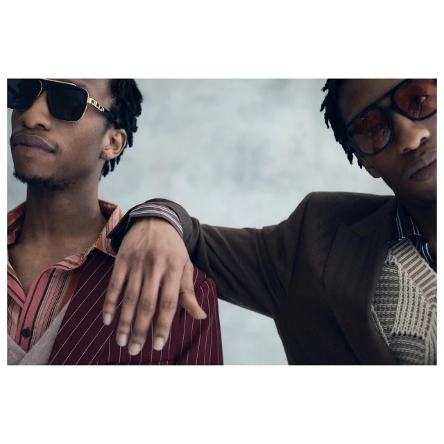 Two stylish African-American men pose indoors wearing fashionable suits and sunglasses, creating a sophisticated and modern portrait. 