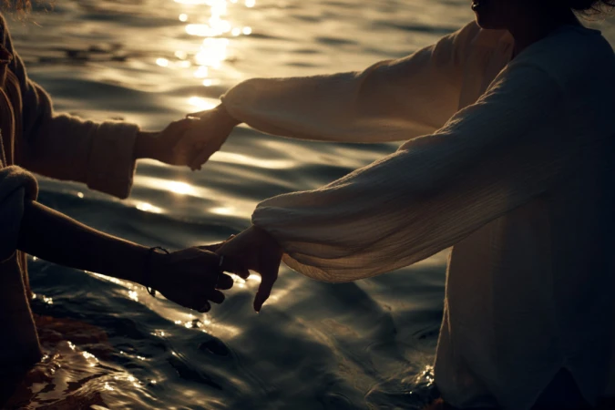 Two people standing waist-deep in water at sunset holding hands while facing each other