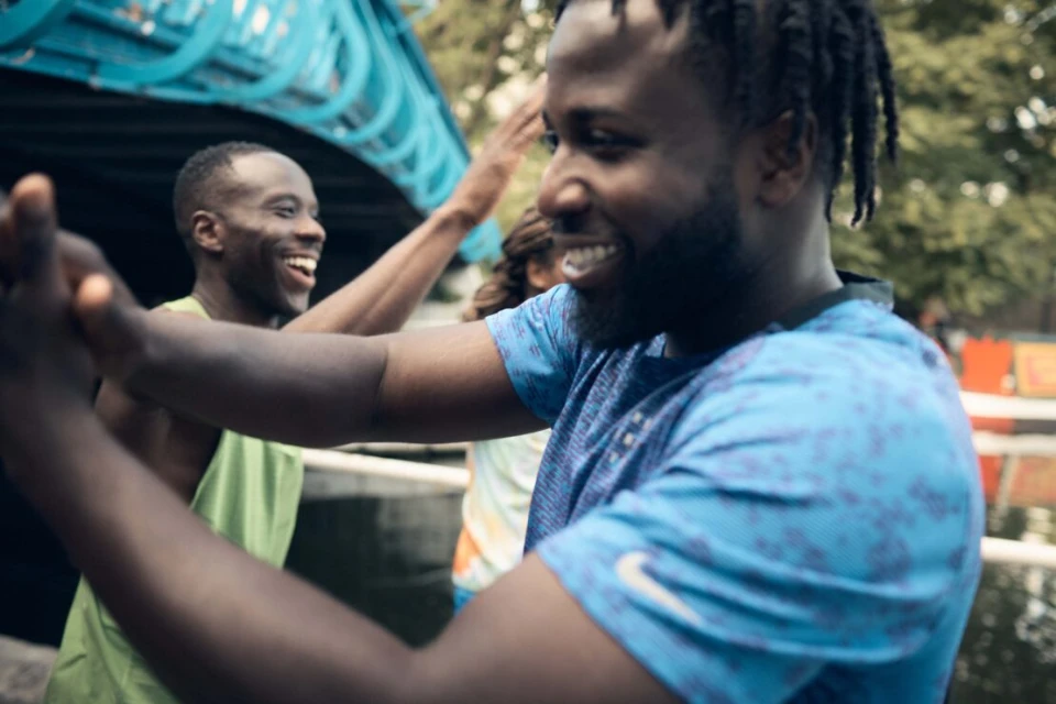 Three adult males with dark complexions are smiling, celebrating, and high-fiving outdoors against a blurred background of trees and a structure.