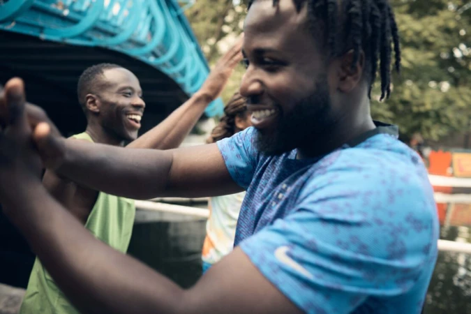 Three adult males with dark complexions are smiling, celebrating, and high-fiving outdoors against a blurred background of trees and a structure.