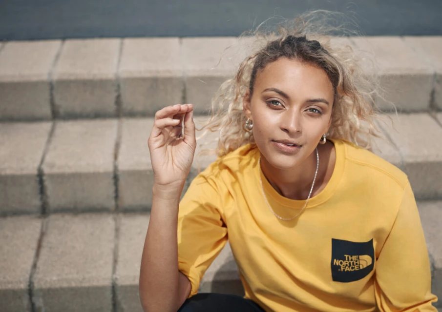 Blonde woman wearing a yellow shirt sitting on concrete steps outdoors. She has silver earrings and a silver necklace.