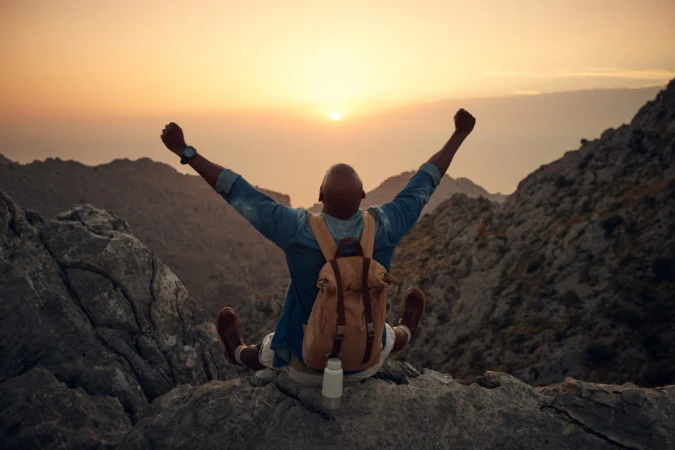 A man with a backpack sitting on a rocky peak with arms raised against a sunset sky by photographer Olly Burn