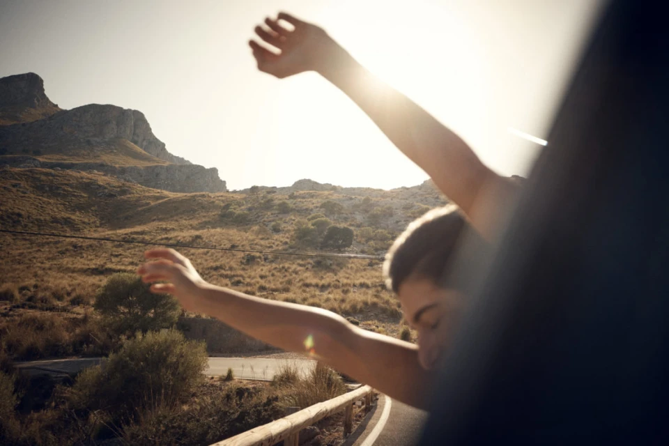 Person in a car stretches arms out the window against a sunlit mountain backdrop. Road trip and freedom concept with blue car interior.