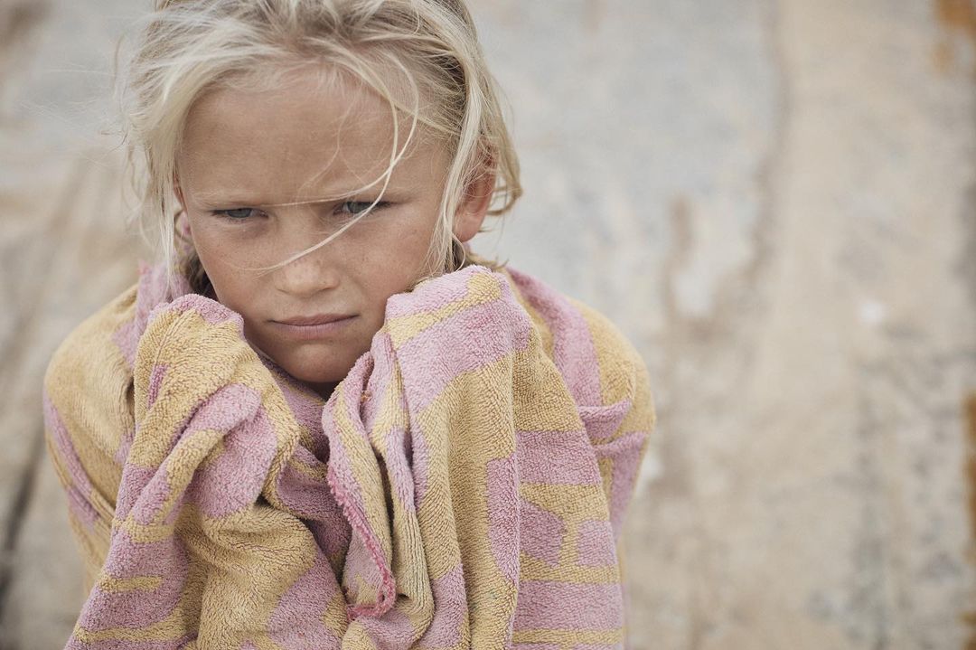 Close-up portrait of a fair-skinned child wrapped in a striped towel, looking slightly downward against a neutral, textured background.