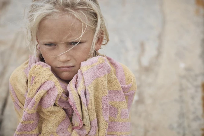 Close-up portrait of a fair-skinned child wrapped in a striped towel, looking slightly downward against a neutral, textured background.