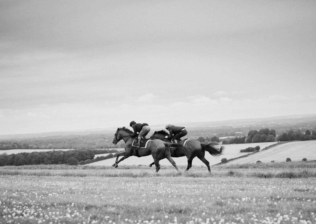 Black and white image of two adult riders on horses galloping in a field with rural landscape background on a cloudy day.