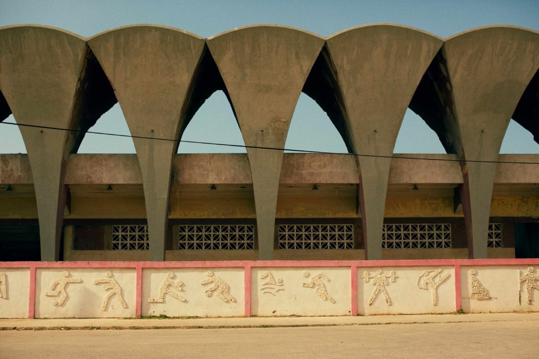 Exterior shot of a concrete sports stadium with arched supports and a wall featuring bas-relief figures depicting various sports activities.