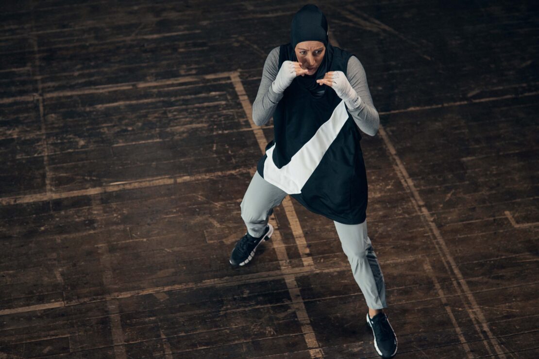 Sporty woman in a hijab, grey tracksuit, and boxing hand wraps in a defensive boxing pose on a wooden floor under even lighting.