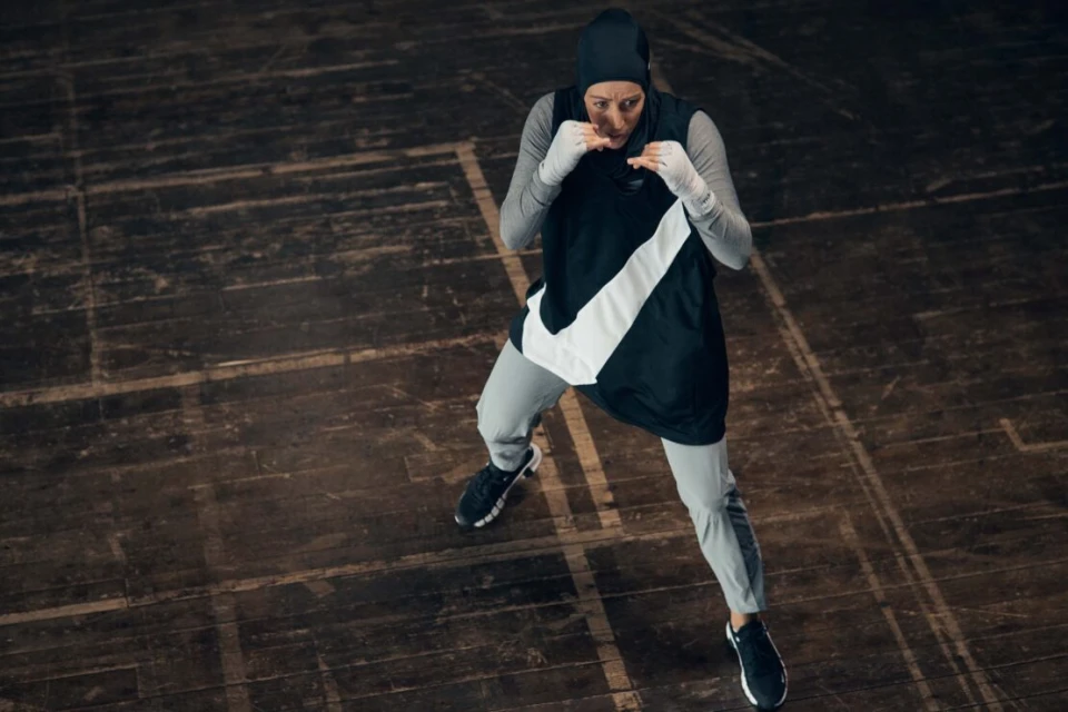 Sporty woman in a hijab, grey tracksuit, and boxing hand wraps in a defensive boxing pose on a wooden floor under even lighting.